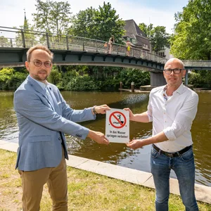 Bezirksamtsleiter Michael Werner-Boelz (Grüne, r.) und Kai-Uwe Jacobsen vom Fachamt Management des öffentlicher Raums stellen die neuen Schilder vor. Im Hintergrund springen Jugendliche von der Brücke ins Wasser.