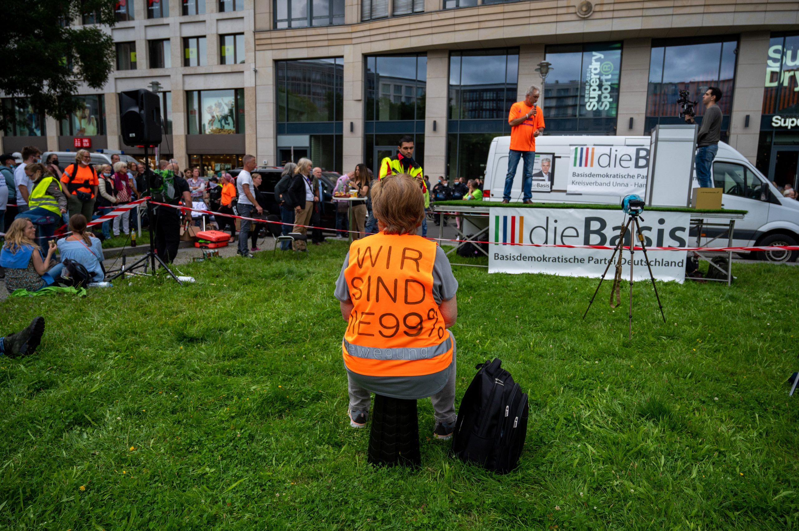 Ein Demonstrant gegen die Corona-Politik der Regierung mit einer Warnweste "Wir sind die 99 Prozent" sitzt auf einer Wiese am Leipziger Platz und hört einem Vertreter der Partei Die Basis zu.