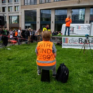 Ein Demonstrant gegen die Corona-Politik der Regierung mit einer Warnweste "Wir sind die 99 Prozent" sitzt auf einer Wiese am Leipziger Platz und hört einem Vertreter der Partei Die Basis zu.