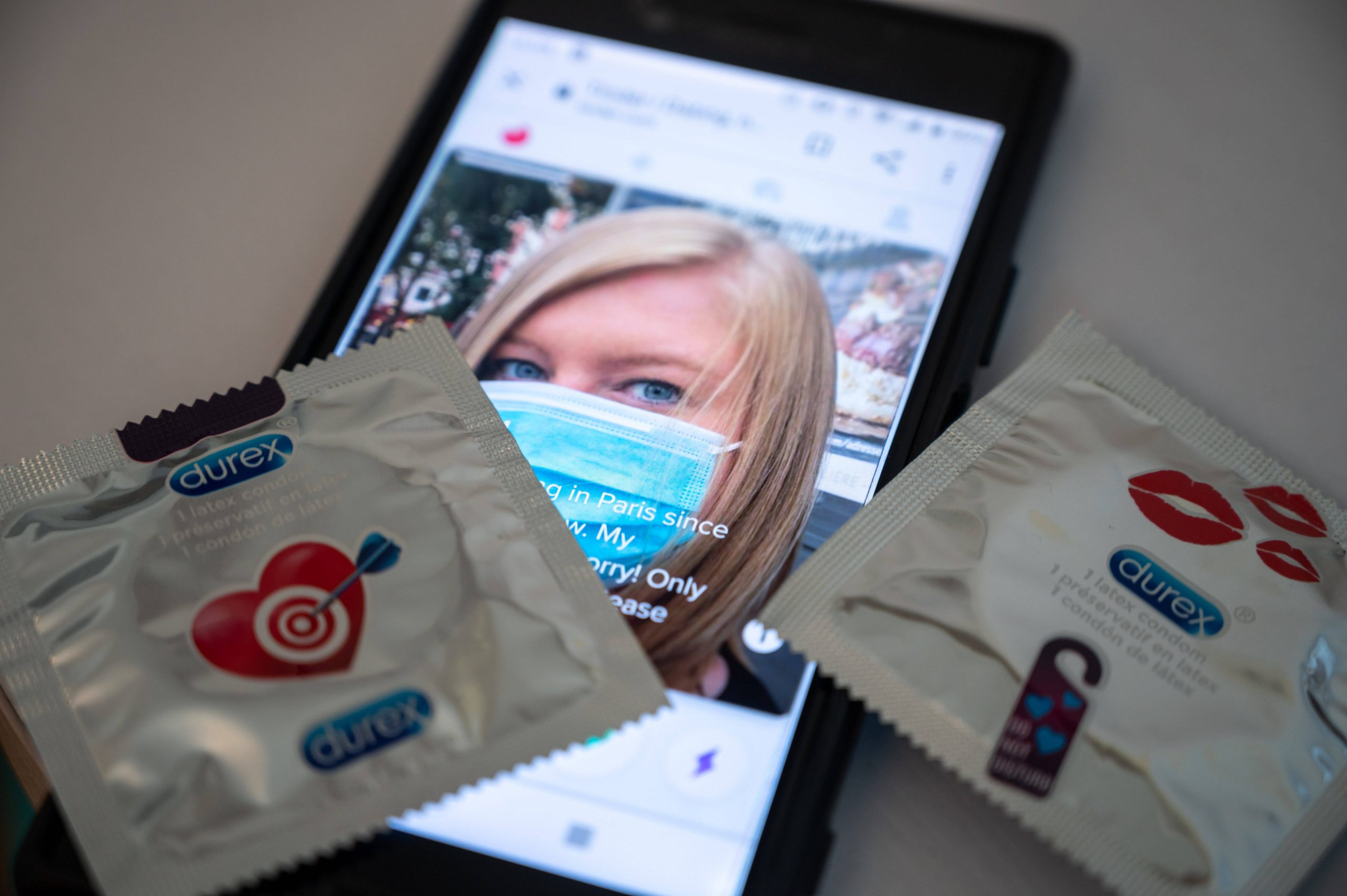 Ein Smartphone auf dem zwei Kondome liegen. Der Bildschirm zeigt das Dating-Profil einer Frau mit Maske.