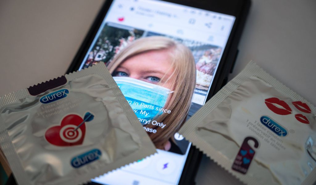 Ein Smartphone auf dem zwei Kondome liegen. Der Bildschirm zeigt das Dating-Profil einer Frau mit Maske.