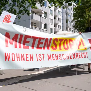 Demonstrationsteilnehmer stehen mit einem Plakat mit der Aufschrift "Mietenstopp - Wohnen ist Menschenrecht" vor einem Hochhaus.