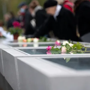 Menschen stehen an Gedenktafeln, auf denen Rosen liegen