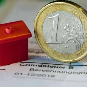Eine Euro-Geldmünze sowie ein Spielzeughaus stehen auf einem Abgabenbescheid für die Entrichtung der Grundsteuer.