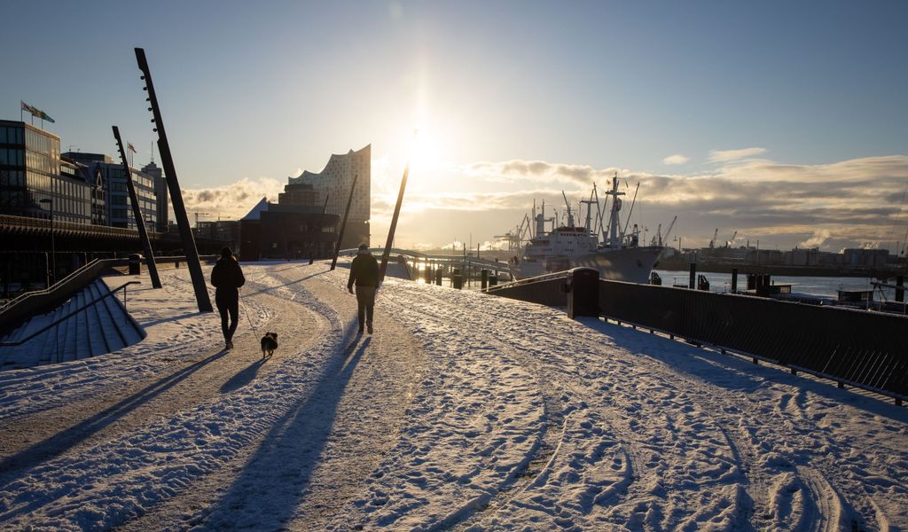 Passanten mit Hund sind auf der verschneiten Elbpromenade bei strahlendem Sonnenschein an den Landungsbrücken im Hafen unterwegs. Im Hintergrund ist die markante Silhouette der Elbphilharmonie in der Hafencity zu sehen.