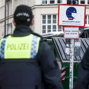 Polizisten auf Streife gehen an einem Schild mit der Aufschrift "Maskenpflicht. Cover your mouth and nose!"