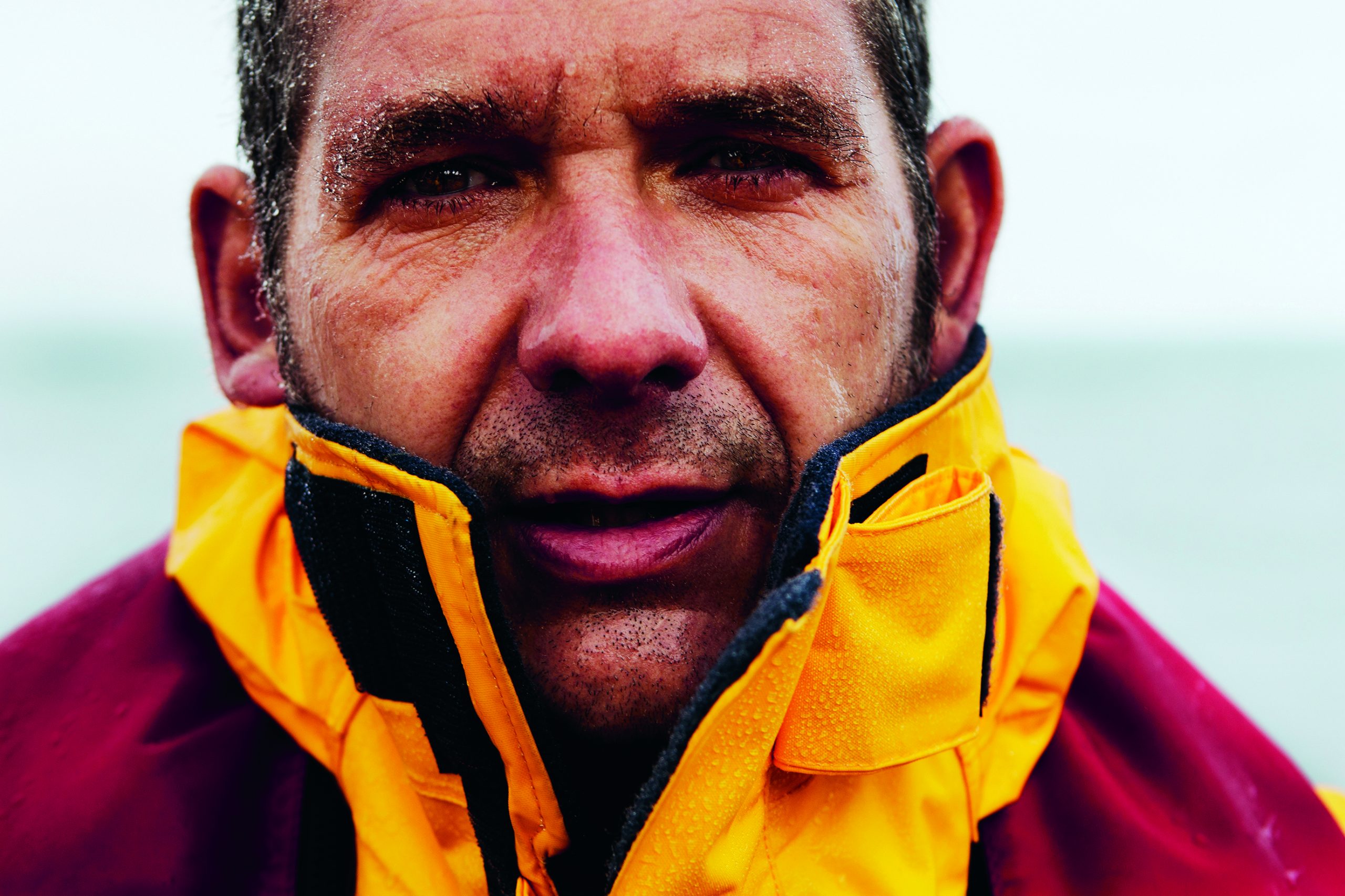 Seenotretter Mark Criddle mit Wasser im Gesicht draußen am Meer, er hat seine Jacke bis zu den Ohren hochgezogen