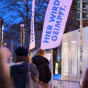 Die Aufschrift "Hier wird geimpft" ist auf einem Aufsteller zu lesen, während mehrere Menschen in einer Schlange vor dem als Impfstation und Schnelltest-Center eingerichteten Container stehen.