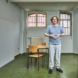 Olli Dittrich steht als Sandro Zahlemann in einem tristen U-Haft-Raum macht mit den Händen die Merkel-Raute