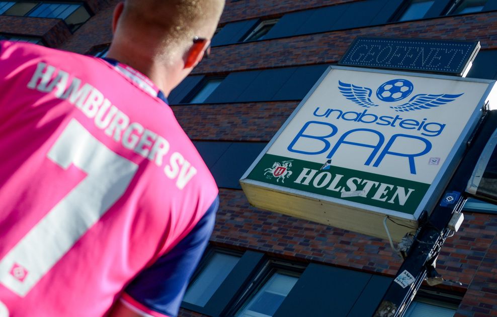 Ein Fan des Hamburger SV steht in der Nähe des Stadions vor einer Bar mit dem Namen "unabsteigbar"