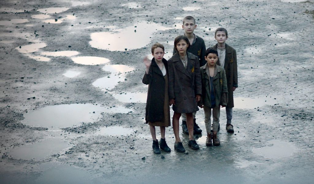 5 Kinder stehen mit KZ-Kleidung und kurzgeschorenen Haaren auf einem Platz voller Pfützen