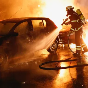 Ein Feuerwehrmann löscht ein brennendes Auto, Flammen ziehen die Straße entlang