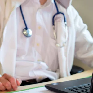Ein Hausarzt sitzt während einer Videosprechstunde in seiner Praxis vor einem Laptop