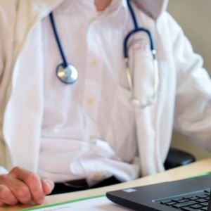 Ein Hausarzt sitzt während einer Videosprechstunde in seiner Praxis vor einem Laptop