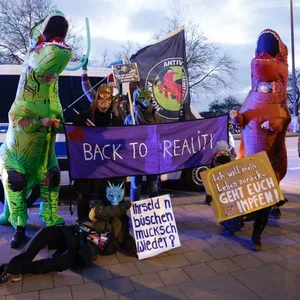 Demonstrierende in Dinosaurier-Kostümen posieren mit Schildern wir "Back to reality"