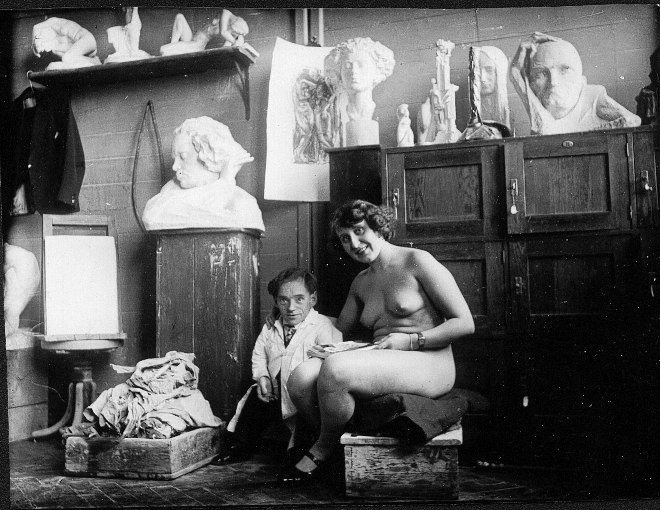 Der Künstler steht neben seinem sitzenden Aktmodell im Atelier, um die beiden herum Büsten auf Regalen und Schränken