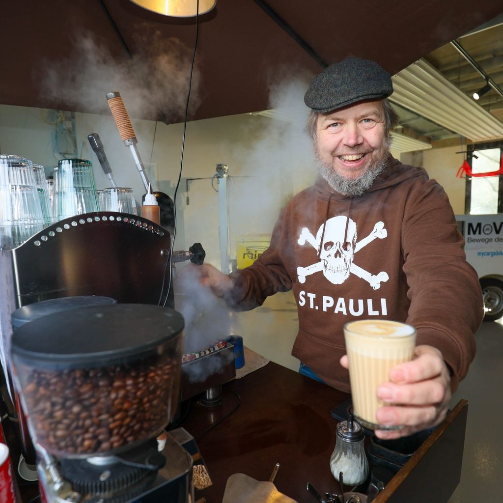 Ein Mann hält einen frisch zubereiteten Kaffee in die Kamera und lächelt