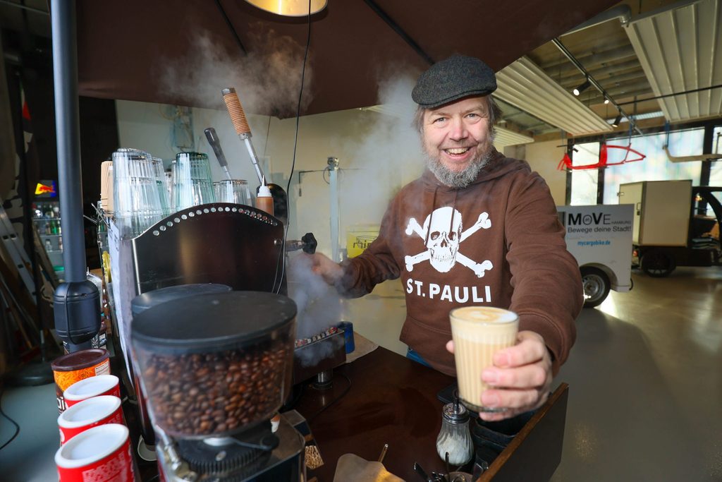 Ein Mann hält einen frisch zubereiteten Kaffee in die Kamera und lächelt