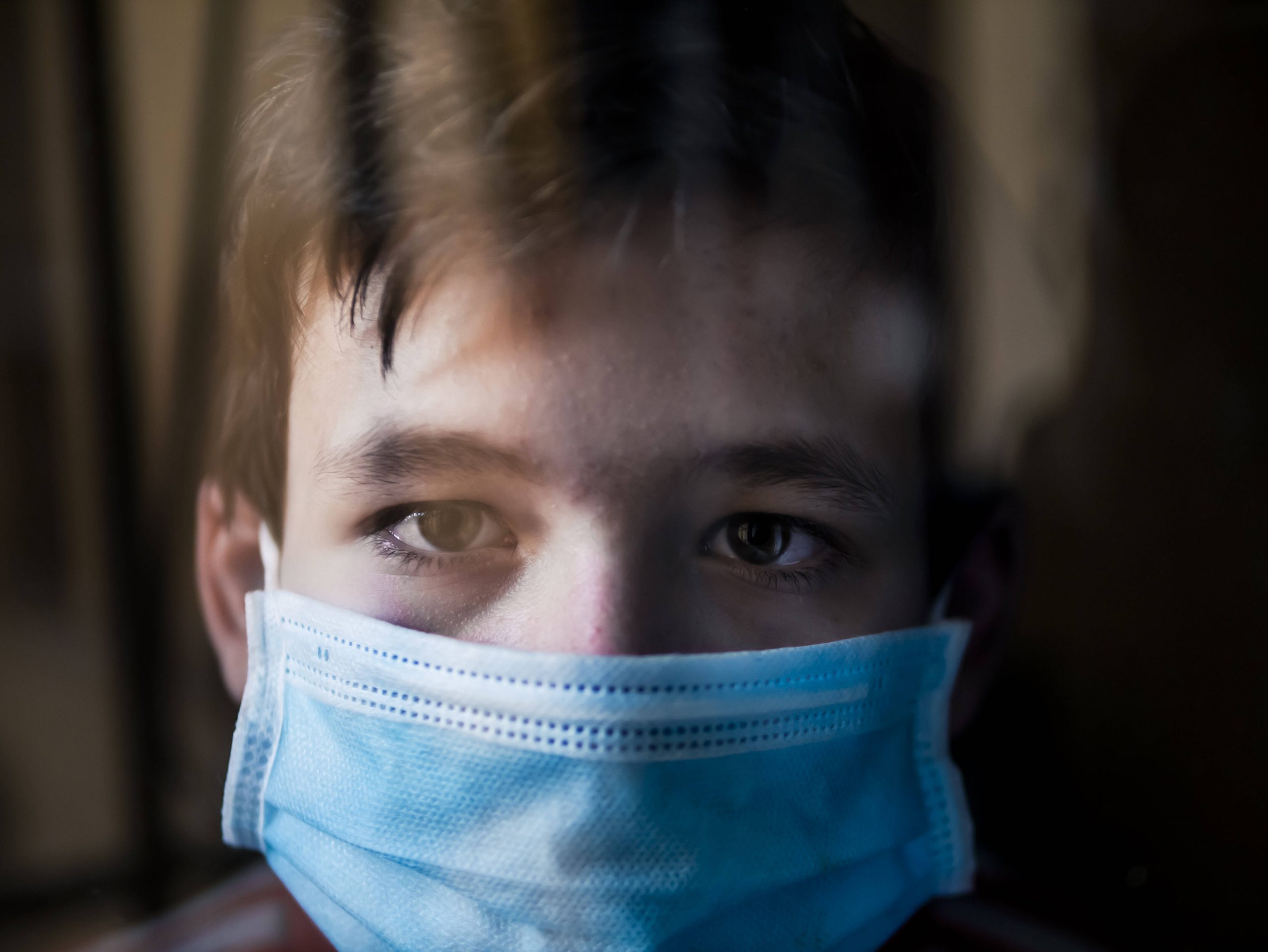 Trauriger Junge mit medizinischer Gesichtsmaske hinter einem Fenster.
