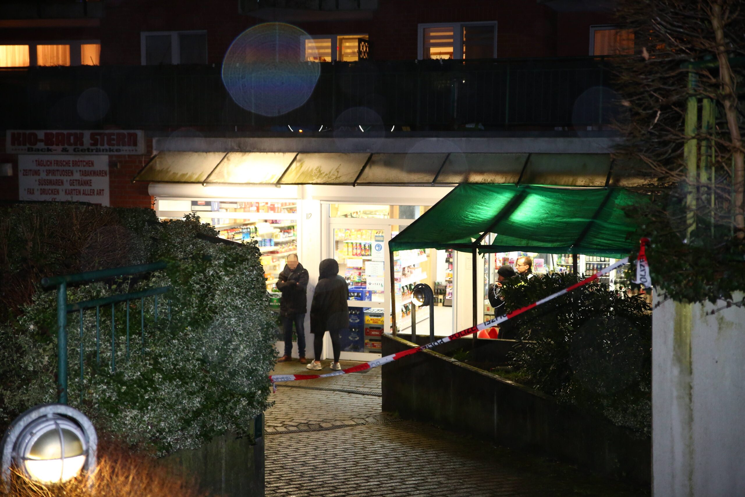 Rot-weißes Flatterband ist vor einem Kiosk angebracht, Männer und Polizisten stehen vor der Tür