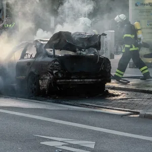 Feuerwehrkräfte löschen einen brennenden Mercedes, daneben steht ein Mann in gelber Jacke