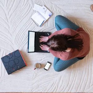 Eine Frau sitzt auf dem Teppich im Homeoffice und arbeitet an einem Laptop.