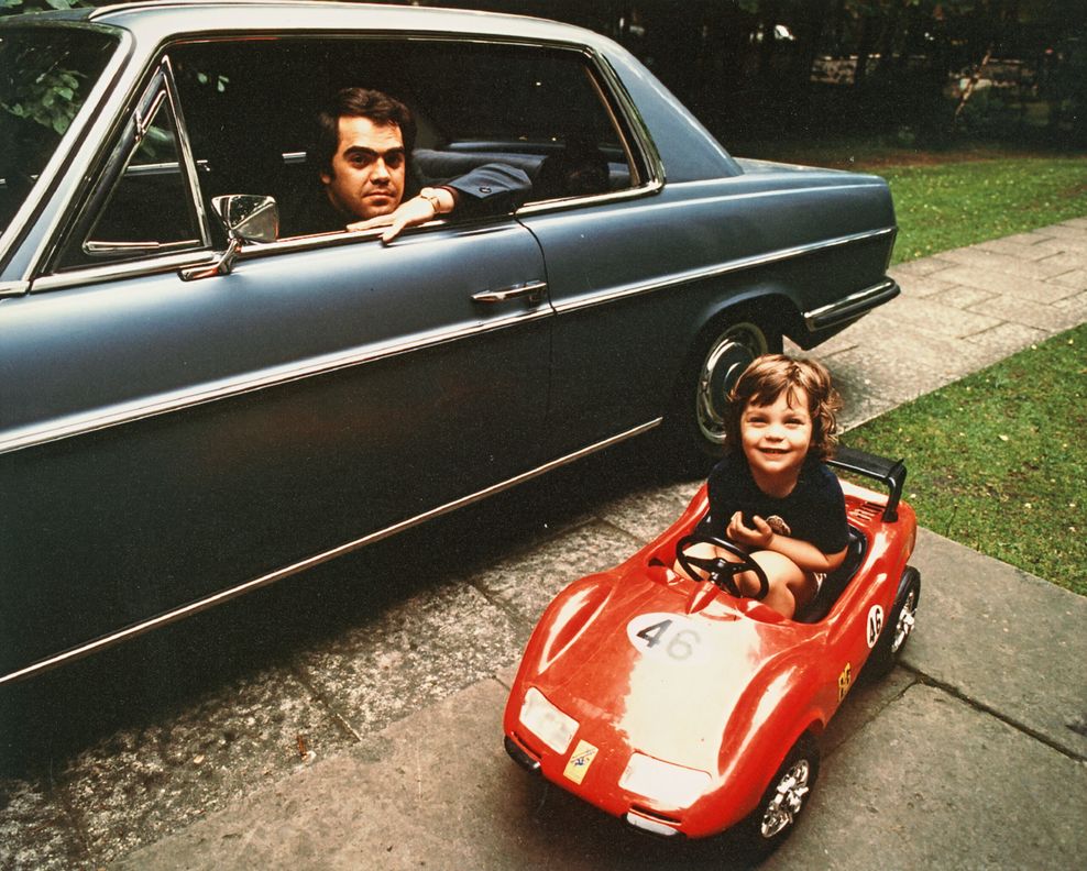 Eugen Cicero sitzt in seinem Auto und schaut aus dem Wagenfenster auf sein Kind Roger, der Junge sitzt in einem roten Kinderauto
