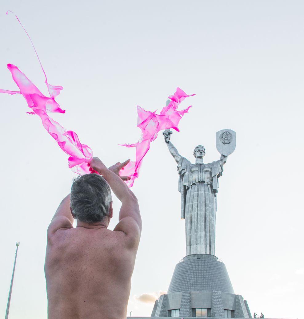 Von hinten sieht man einen Mann mit nacktem Oberkörper, der vor einer Statue steht und pinken Stoff rumwirbelt.