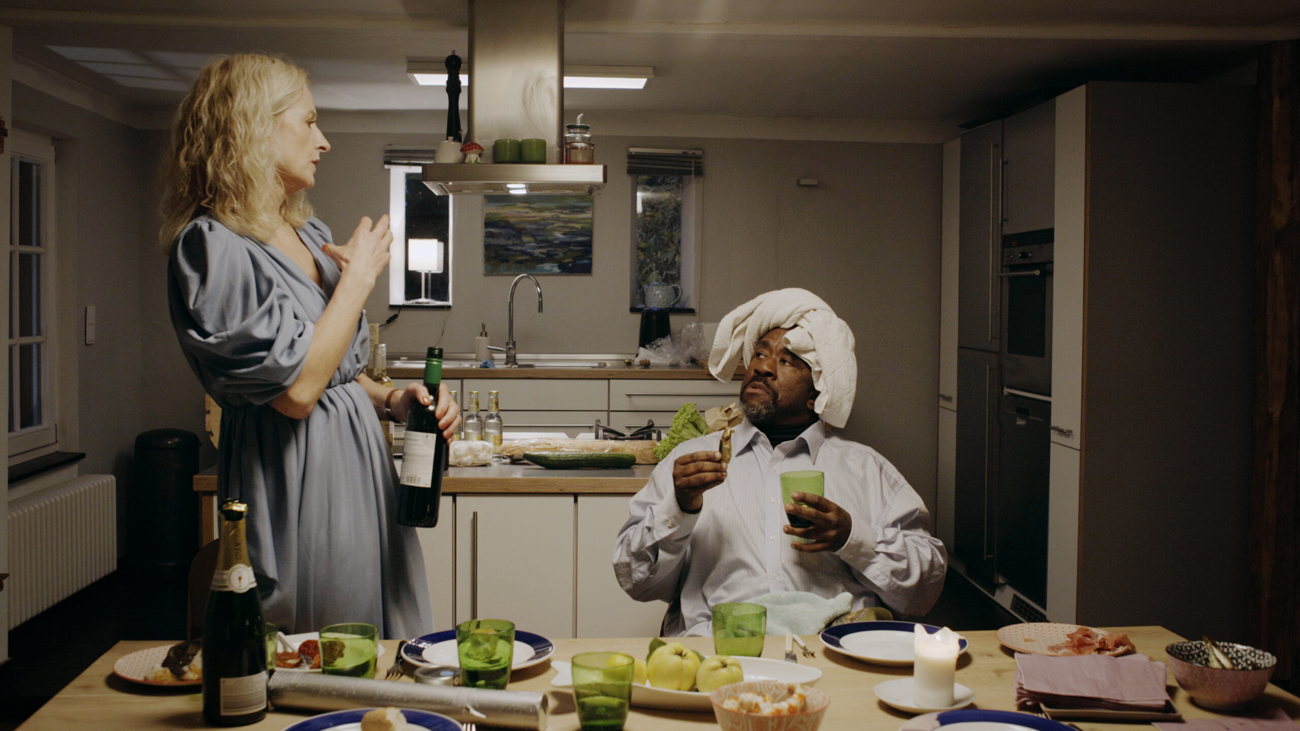 Voller Esstisch in der Küche, eine Frau steht links mit einer Flasche Wein, rechts sitzt ein Mann und sieht sie an