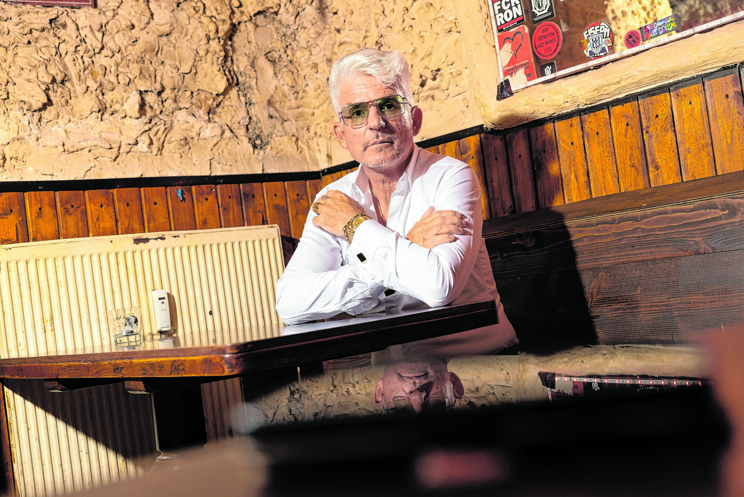 Heinz Strunk sitzt auf einer Kneipenbank am Tisch, trägt Sonnenbrille und blickt in die Kamera