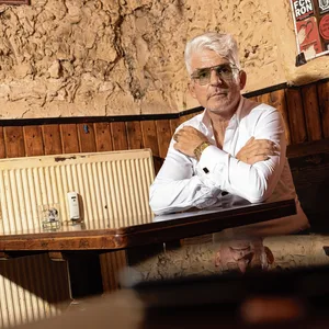 Heinz Strunk sitzt auf einer Kneipenbank am Tisch, trägt Sonnenbrille und blickt in die Kamera