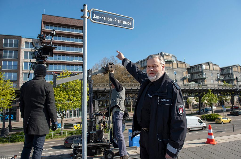 Ein Schauspieler in Polizei-Uniform zeigt auf das Straßenschild.