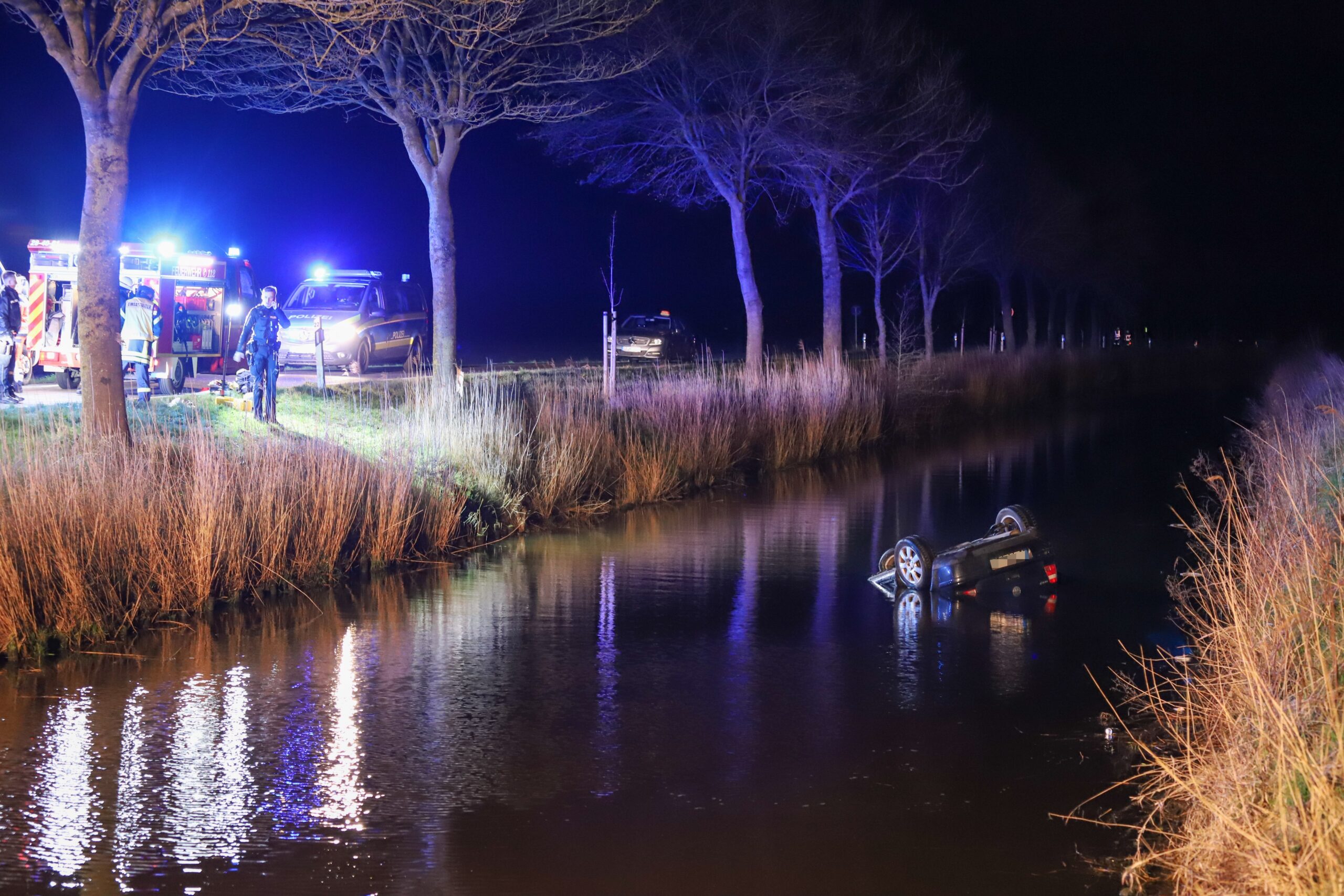 An einer Landstraße stehen Polizei- und Rettungswagen, daneben im Wassergraben liegt kopfüber ein Auto