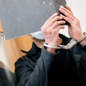Der Angeklagte steht Prozessbeginn mit Handfesseln in einem Saal im Landgericht und bedeckt sein Gesicht mit einem Aktenordner