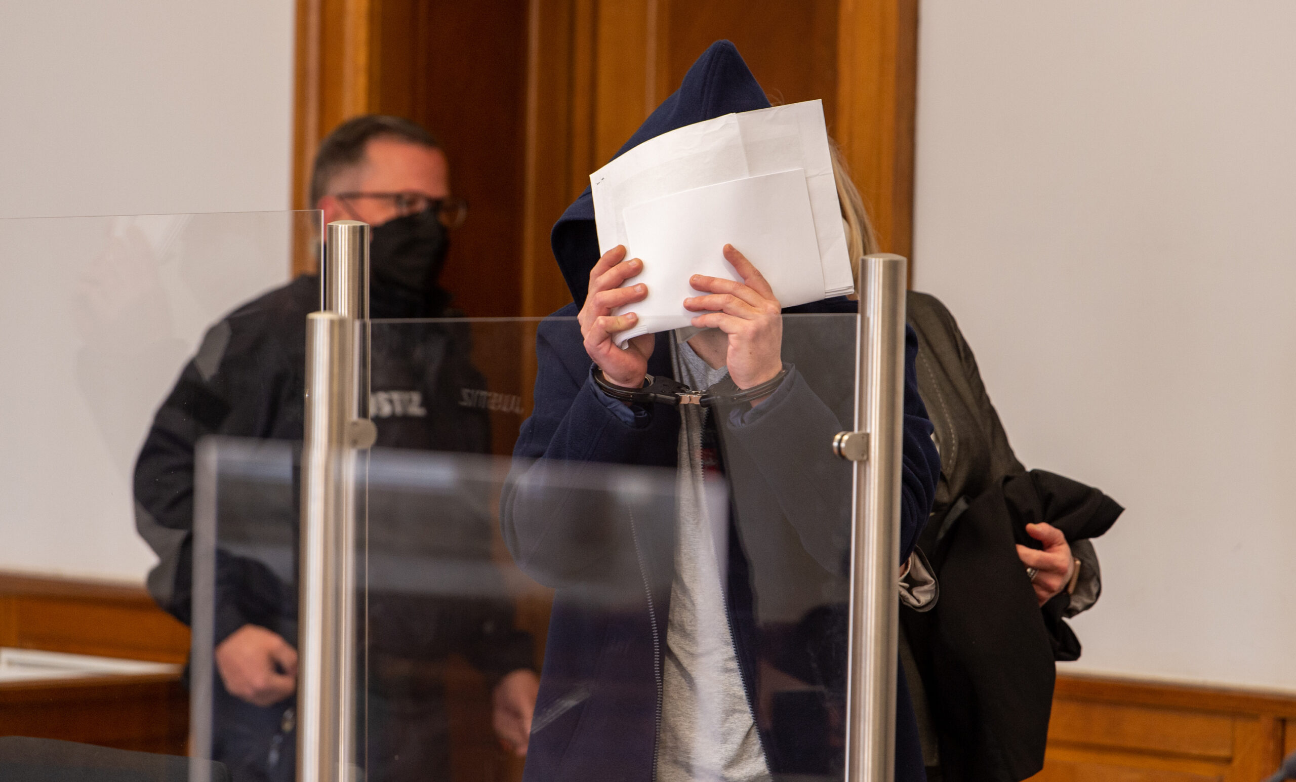 Der Angeklagte wird in den Saal vom Landgericht geführt und verdeckt sein Gesicht. Dem 38-Jährigen wird vorgeworfen, im Oktober 2021 in Lüneburg seine im 7. Monat schwangere Lebensgefährtin durch Messerstiche getötet zu haben.