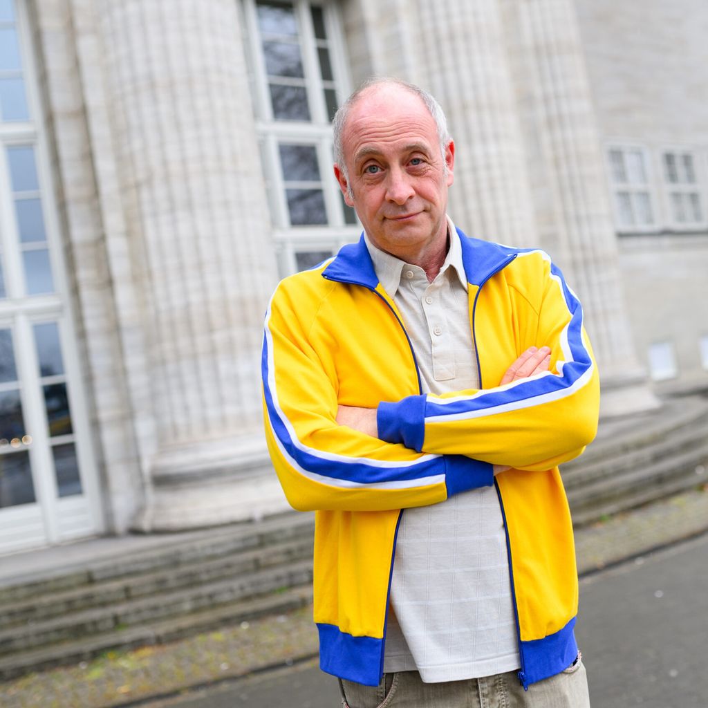 Kabarettist Alfons steht mit verschränkten Armen und in gelb-blauer Jacke