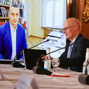 Drei Personen sitzen an Tischen mit Mikrofonen, Klitschko ist per Video zugeschaltet