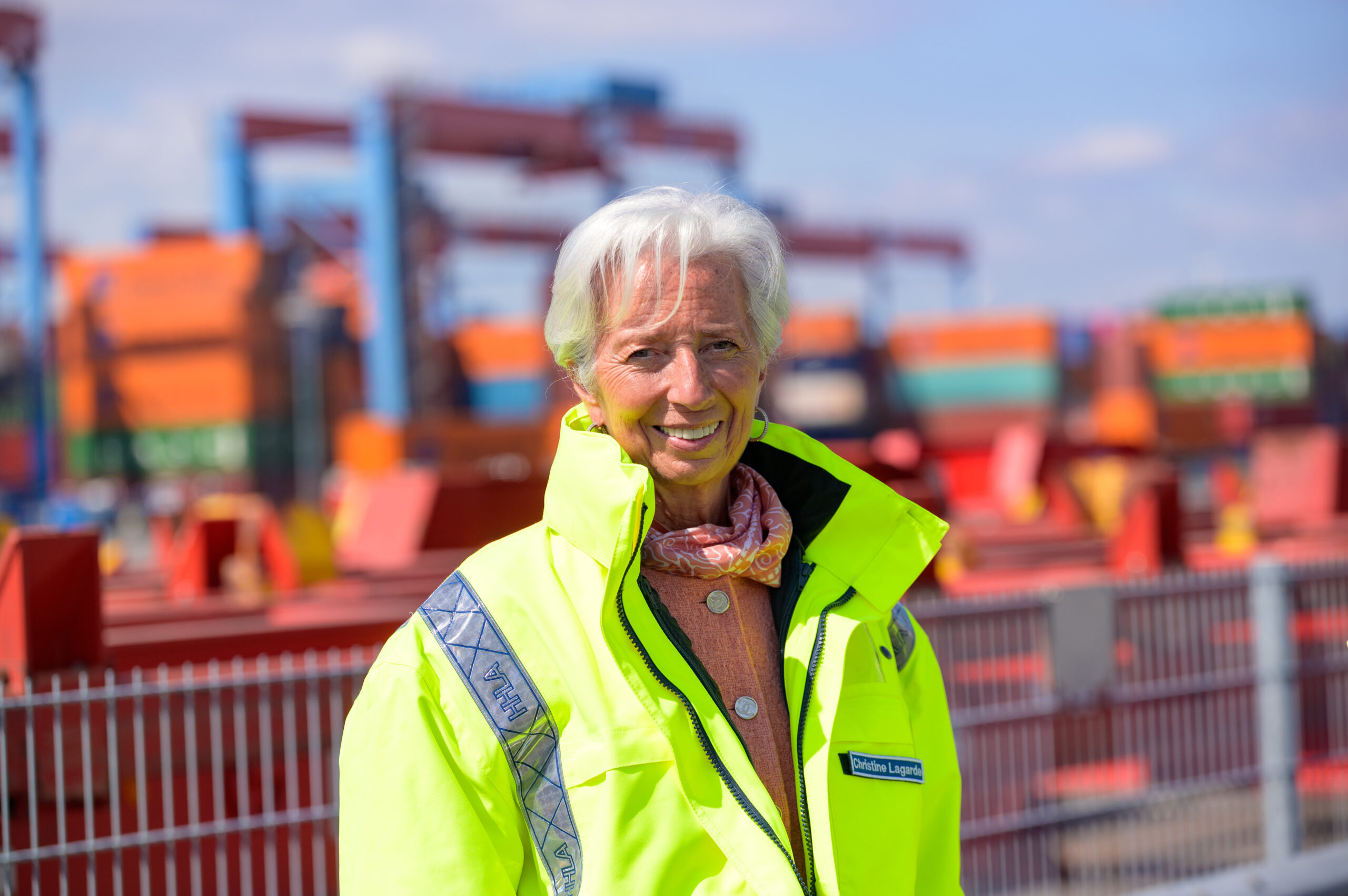 Christine Lagarde steht in gelber Jacke vor Kränen und Containern