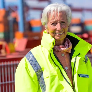 Christine Lagarde steht in gelber Jacke vor Kränen und Containern