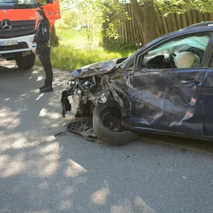 Ein blauer VW steht mit zerstörter Vorderseite auf einer Straße, daneben Feuerwehrleute