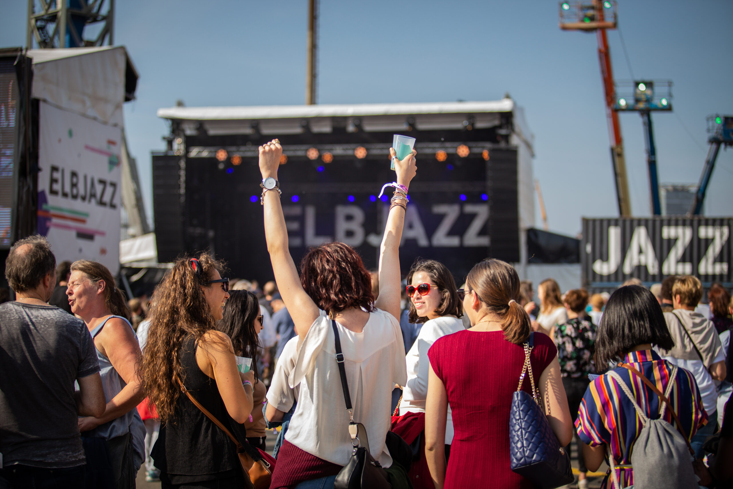 Im Hintergrund eine der Elbjazz-Bühnen, blauer Himmel, im Vordergrund Publikum von hinten, eine Zuschauerin in der Mitte reißt die Arme in die Höhe