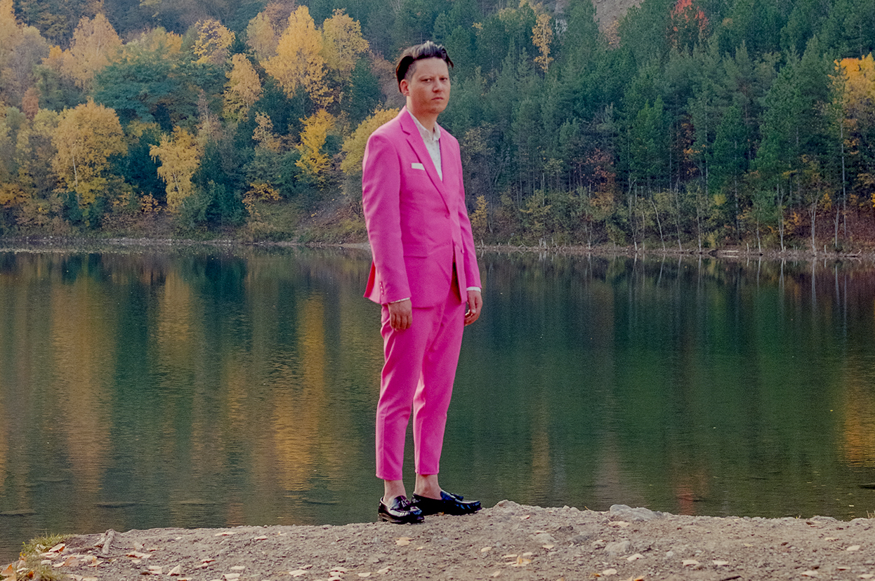 Konstantin Gropper steht an einem See, im Hintergrund ein Wald. Er trinkt einen sehr pinkfarbenen Anzug. Mit Einstecktuch