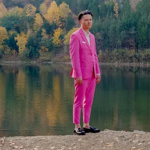 Konstantin Gropper steht an einem See, im Hintergrund ein Wald. Er trinkt einen sehr pinkfarbenen Anzug. Mit Einstecktuch