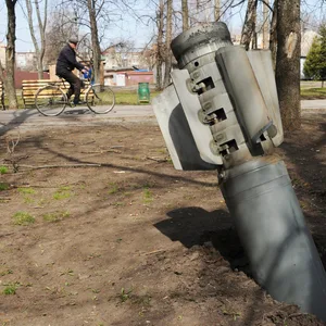 Ein Mann fährt mit dem Fahrrad durch Charkiw, neben ihm ragt das Endstück einer Rakete aus der Erde.