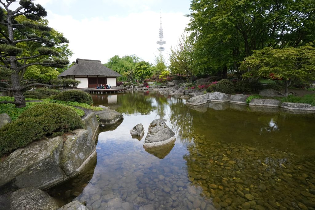 Passanten sitzen auf einem Steg an einem kleinen Teich im Park „Planten un Blomen“.