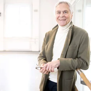 John Neumeier lehnt entspannt an einer Ballettstange und lächelt in die Kamera.