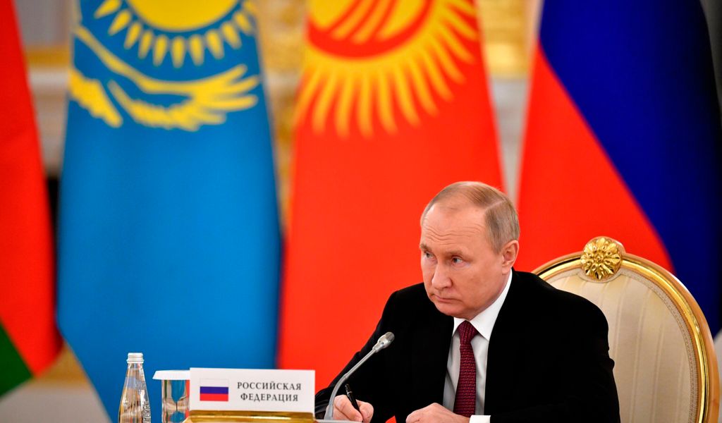 Putin sitzt an einem Tisch, hinter ihm Nationalflaggen