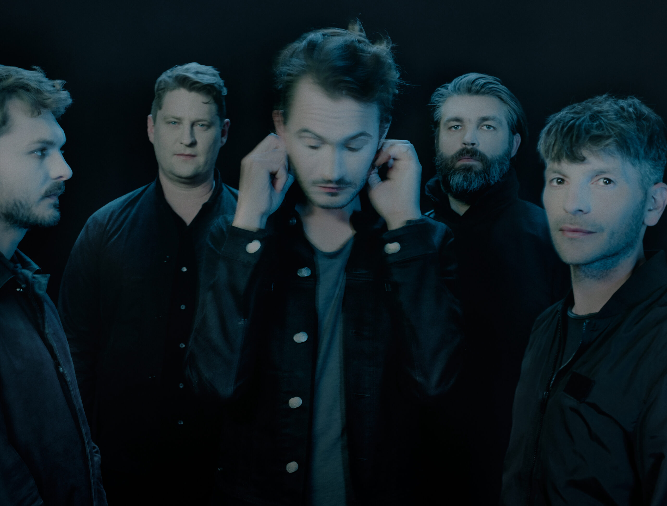 Sehr düsteres Foto, fünf Männer, in der Mitte Tom Smith, der sich an die Ohren fasst und unscharf ist. Der Rest der Band schaut zum Teil in die Kamera