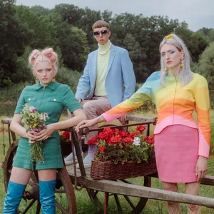 Die 3 Blond-Musiker:innen stehen auf einer Wiese an einer mit Blumen geschmückten Holzkarre. Sie tragen sehr bunte Kleidung