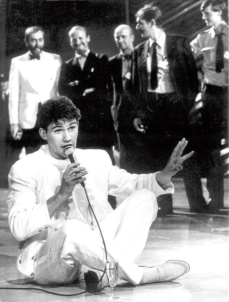 Schwarzweißfoto, Johnny Logan sitzt auf dem Boden im weißen Anzug, Mikrofon in der Hand, vor ihm ein Glas Sekt. Im Hintergrund stehen Männer